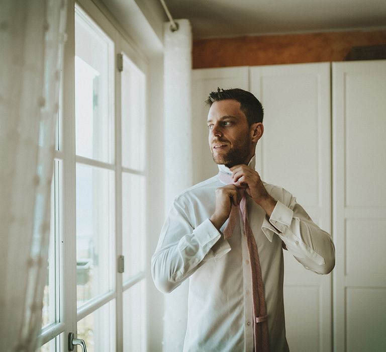 Groom looks out the window on his wedding day as he does up his shirt