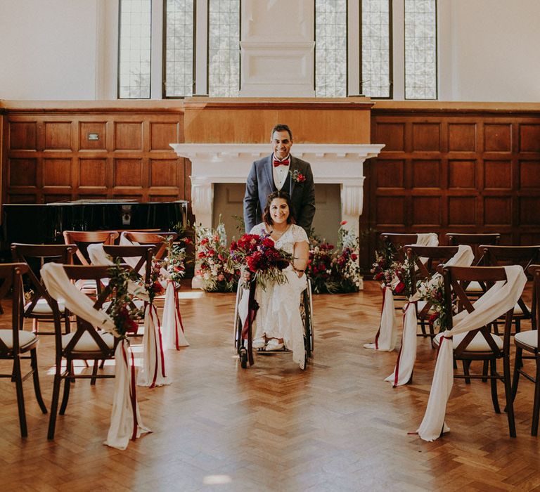 Groom in a blue suit and burgundy bow tie pushing his bride in a wheelchair up the aisle at their Dulwich College wedding with drape and flower arrangements decorating the aisle chairs 