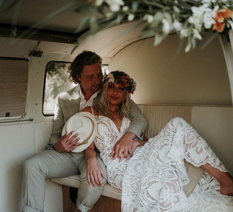 Groom in white shirt and linen suit holding white straw fedora sits in campervan hugging bride in white lace boho wedding dress and tropical flower crown for birds of paradise wedding inspiration