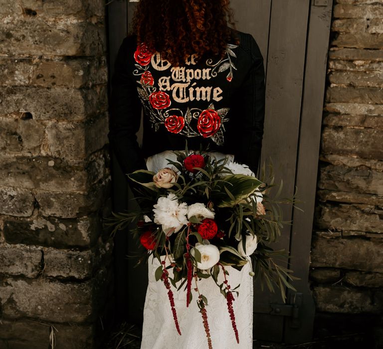 Black bride in a leather jacket with red and gold floral motif holding her bouquet behind her back 