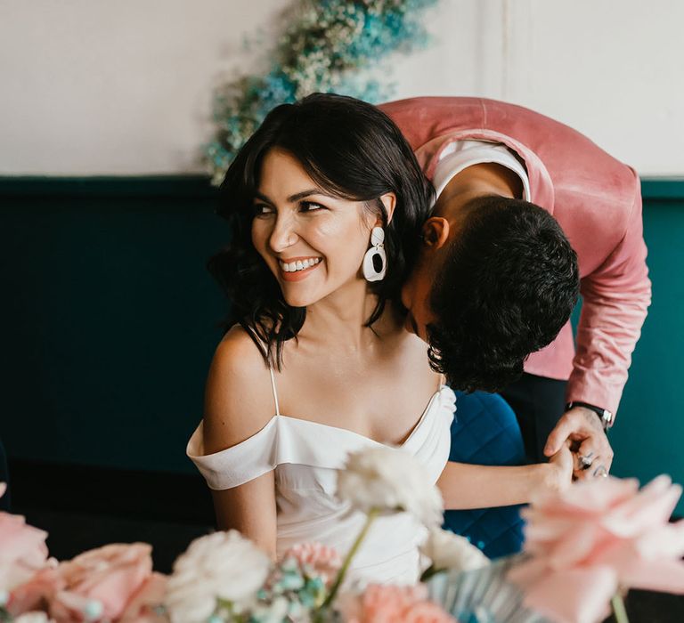 Groom in a pink velvet jacket kissing his bride on the shoulder with short curly hair wearing a cold shoulder wedding dress and dangly earrings 