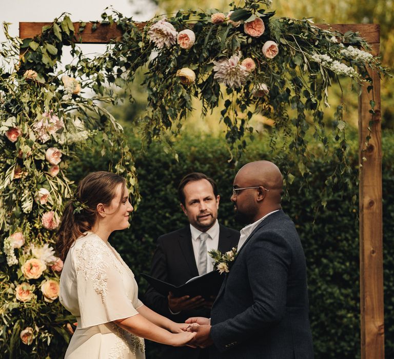 Couple stand under flower arch holding hands as in front of celebrant during marriage ceremony. 