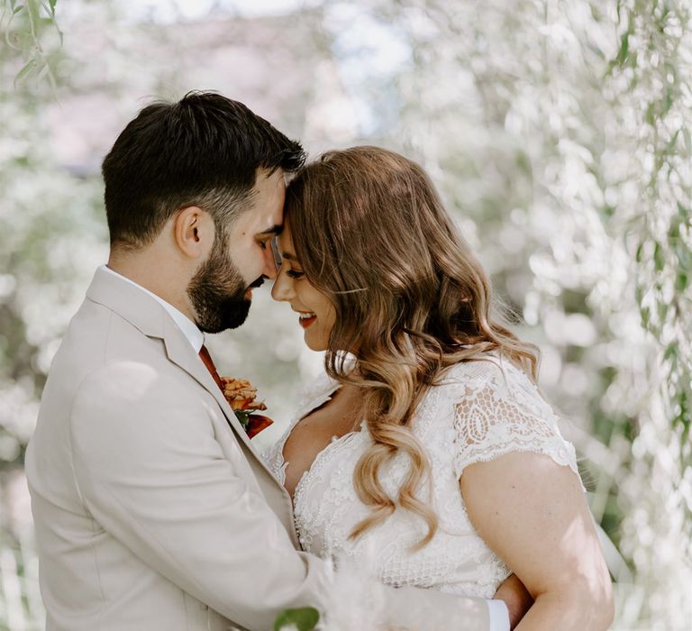 Bride in homemade lace wedding dress with mixed rose bridal bouquet and curled hair hugs groom in beige suit with floral buttonhole at DIY garden wedding ceremony