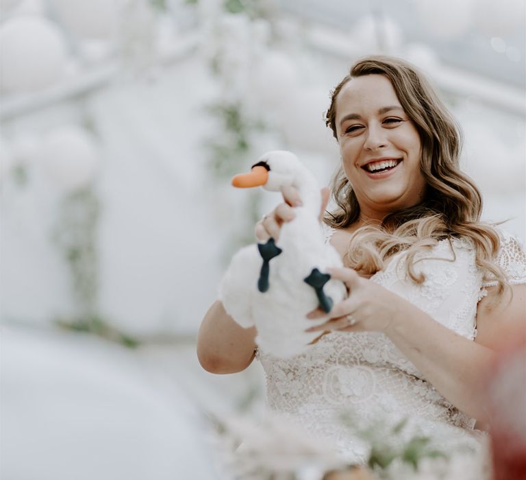 Bride in lace homemade wedding dress and curled hair holding toy swan smiles during marquee wedding reception 