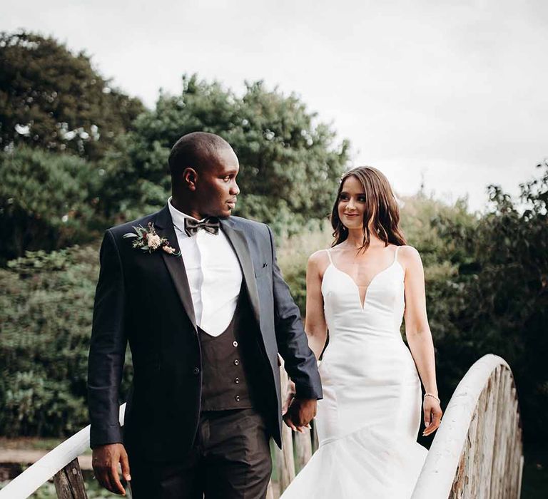 Bride wears love heart shaped Eylsee wedding dress & holds hand with groom in fitted dark suit with a feather & burgundy mixture boutonniere 