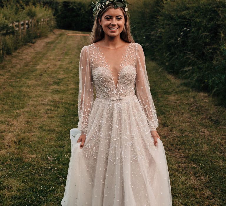 Bride in long mesh sleeve embroidered Jule Vino wedding dress with v neck and boho flower crown walks through field after outdoor wedding reception