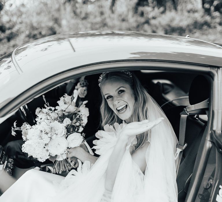 Bride in Halfpenny London dress and veil holding mixed bridal bouquet waves from car before wedding ceremony