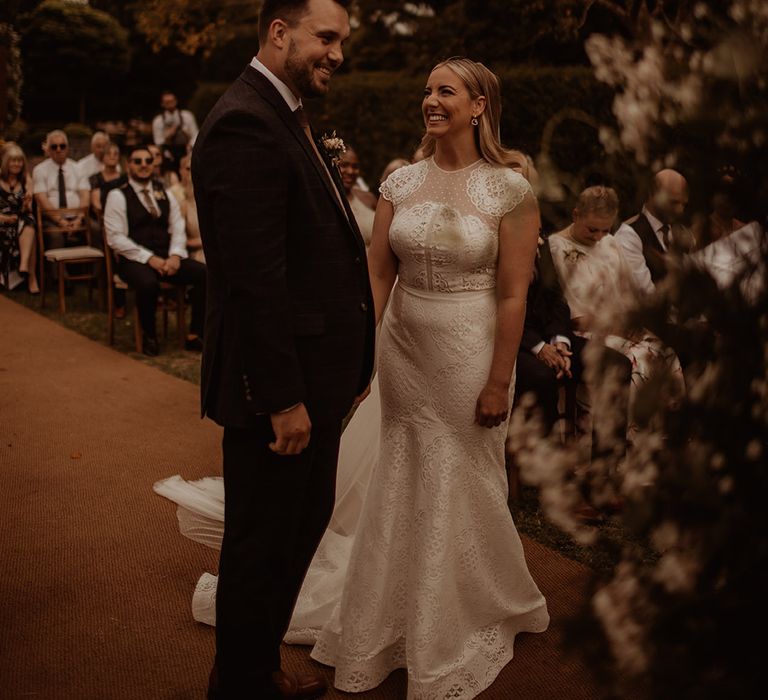 Bride in a Jesus Piero lace fishtail wedding dress with cap sleeves smiling at her groom during their outdoor Dewsall Court wedding 