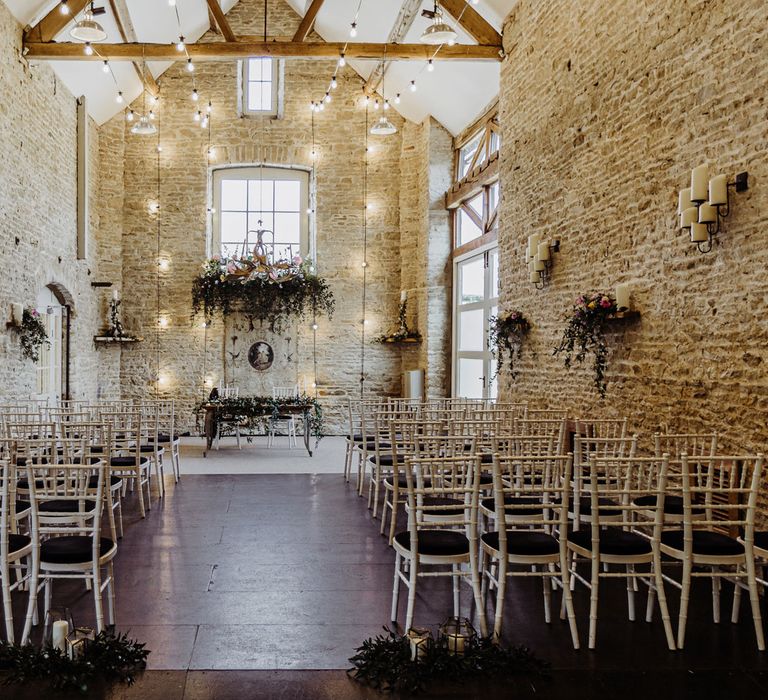 Stone barn wedding ceremony room with Edison light bulbs, bamboo style chairs, candles and foliage