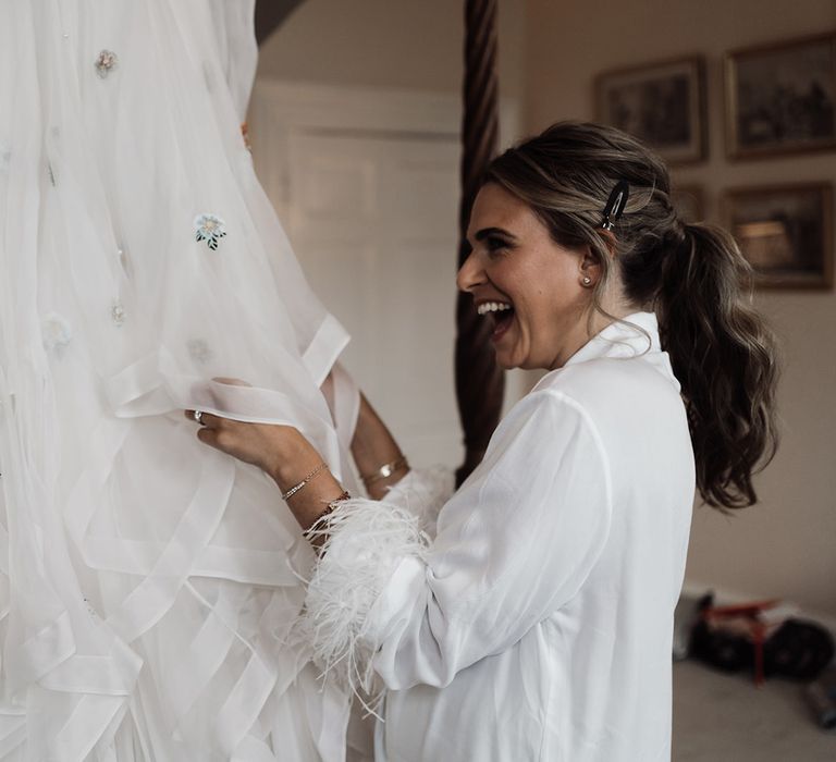 Bride in white fur trim satin robe stands holding train of embroidered Victoria Sanders wedding dress