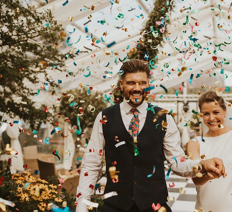 Colourful confetti moment in a clear marquee with groom in a navy waistcoat and floral tie and bride in a slip wedding dress 