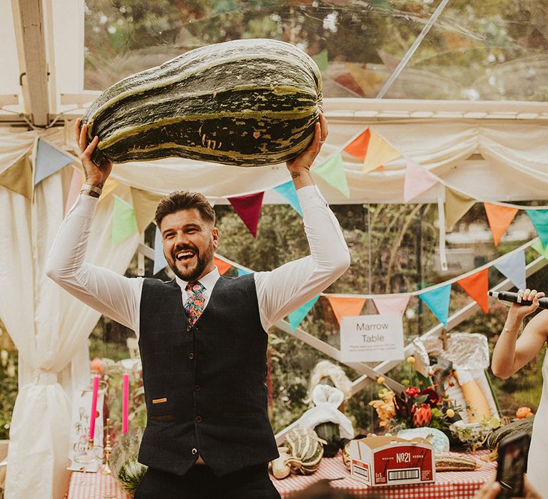 Groom in a navy waistcoat and floral tie holding up the winner of the best in marrow competition