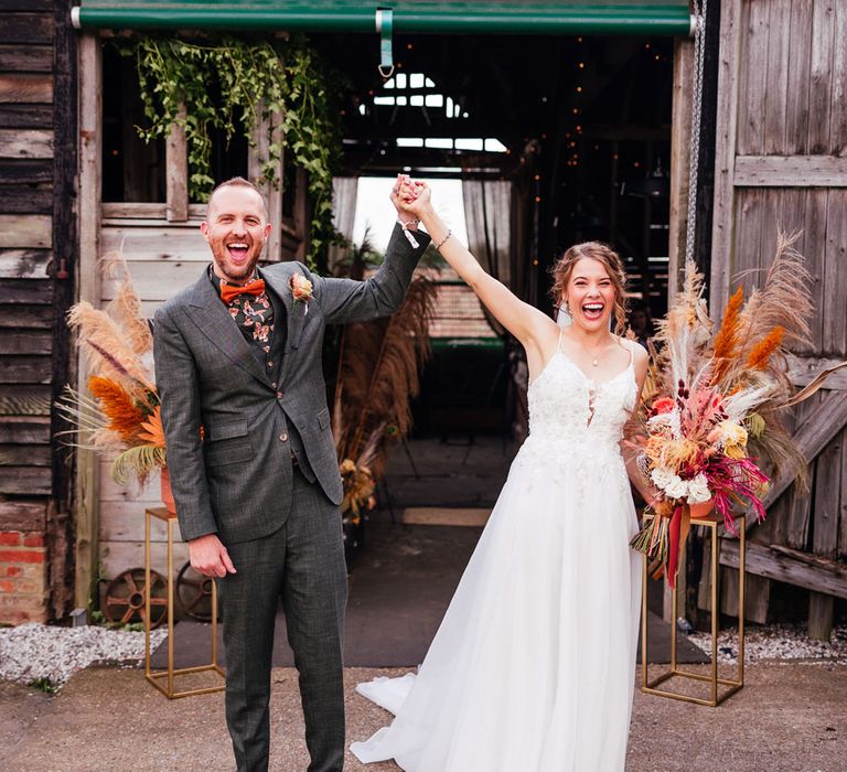 Bride in lace wedding dress with train holding mixed dried floral boho bouquet holds up arm with groom in green three piece suit and pattered shirt as they leave barn wedding ceremony