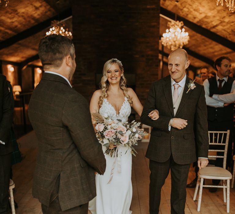 Bride with curled blonde hair and lace wedding dress holding white, green and pink bridal bouquet smiles as she walks arm in arm with father of the bride down barn aisle