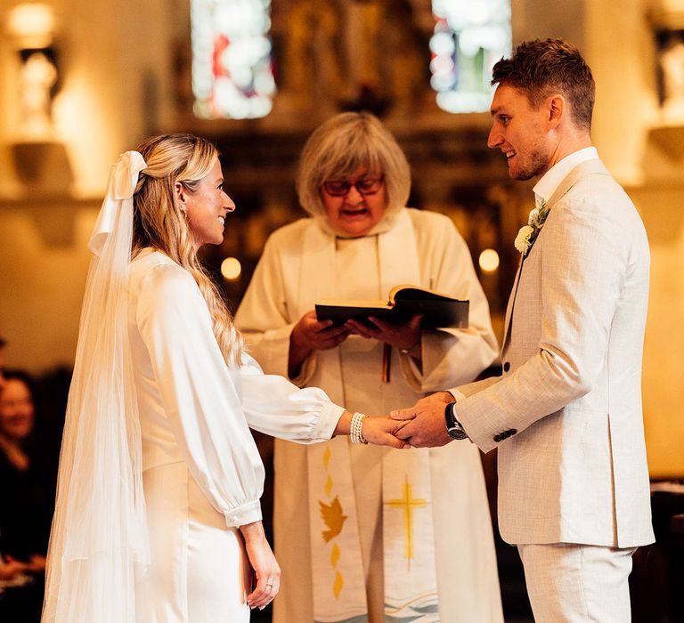 Groom in light grey suit and floral buttonhole holds hand of bride in long sleeve satin wrap top, hair bow and veil during church wedding ceremony