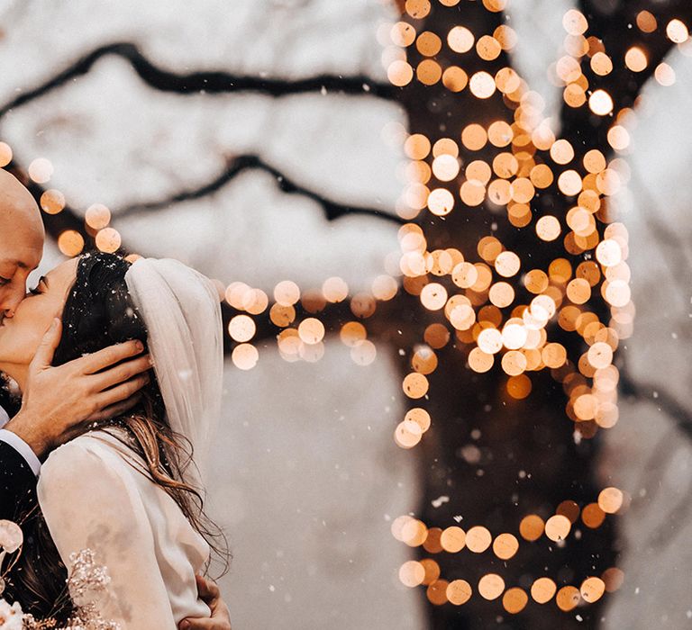 Groom in a black suit kissing his bride in a high neck wedding dress and long wedding veil at their snowy winter wedding 