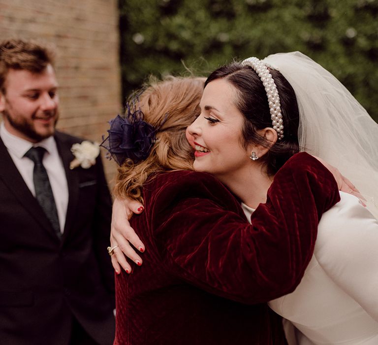 Bride in pearl headband and long veil hugs a wedding guest in red velvet jacket