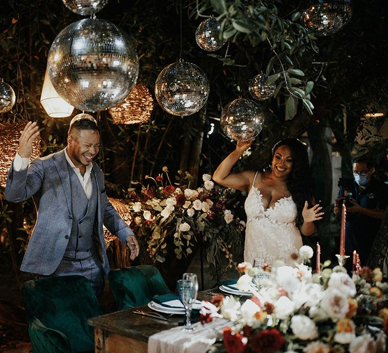 Bride and groom walk with multiple disco balls overhead with velvet green seats and pink candles for private top table 