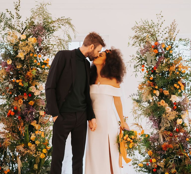 Bride in an off the shoulder wedding dress with side slit and groom in all-black kissing between column golden wedding ceremony flowers