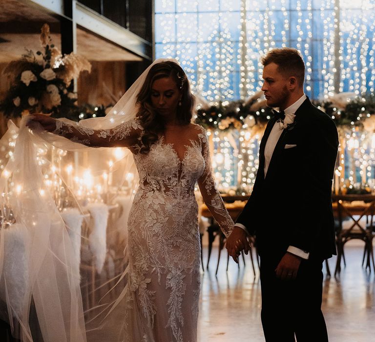 Bride in lace wedding dress holds out her veil as groom looks at her mesmerised in front of fairy lights