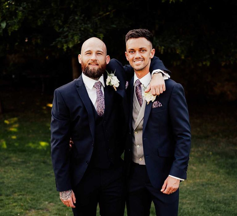 Groom in dark matching suit but with grey waistcoat stands with best man