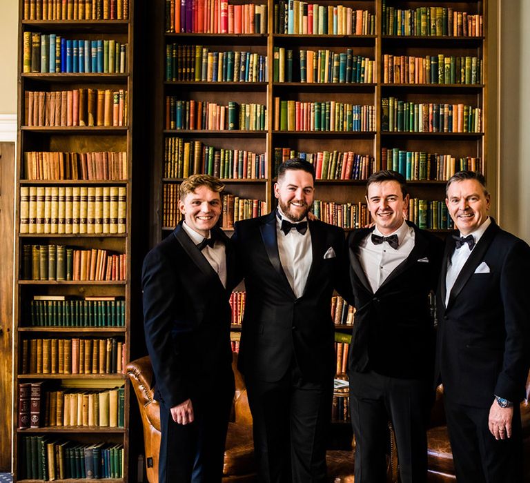 Groom and groomsmen stand in front of bookcase in black tie for autumn wedding 