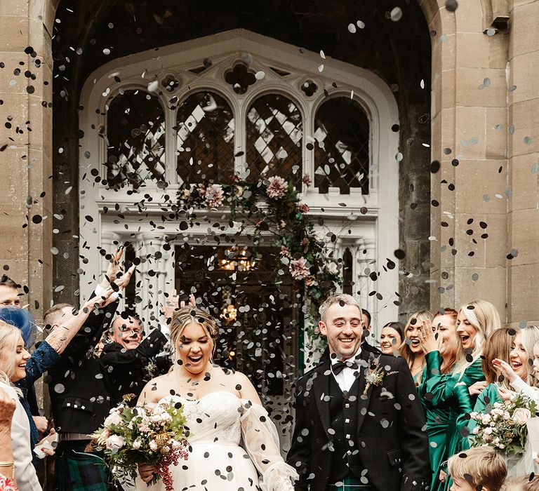 Bride in sheer sleeved off the shoulder wedding dress with groom in black tie and kilt have confetti exit with black circle confetti