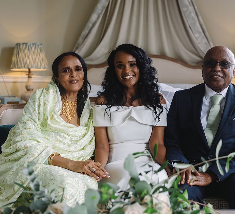 Bride in off the shoulder wedding dress with curly black hair with mother and father in mint green outfits with gold jewellery