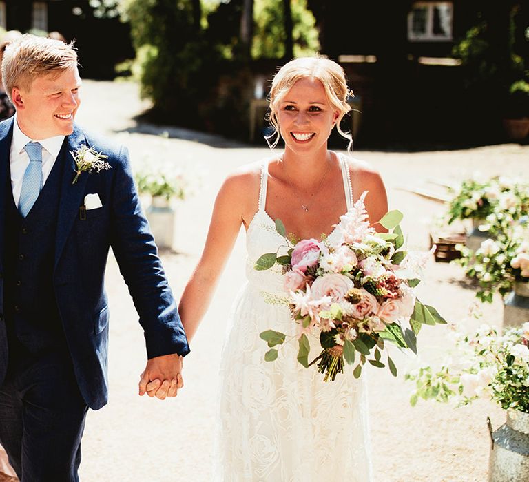 Bride and groom walk hand in hand together after their wedding ceremony with bride in lace boho dress and groom in blue suit 