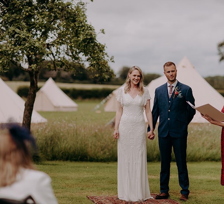 Bride in beaded wedding dress and groom in deep blue suit stand on patterned rug for humanist wedding ceremony 