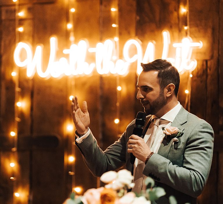 Groom in grey suit jacket with pink tie and pink rose buttonhole gives speech in front of neon 'Worth the Wait' sign 