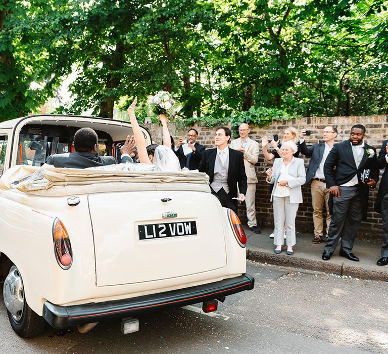 Bride and groom ride off in roof down cream car as wedding guests wave them off 