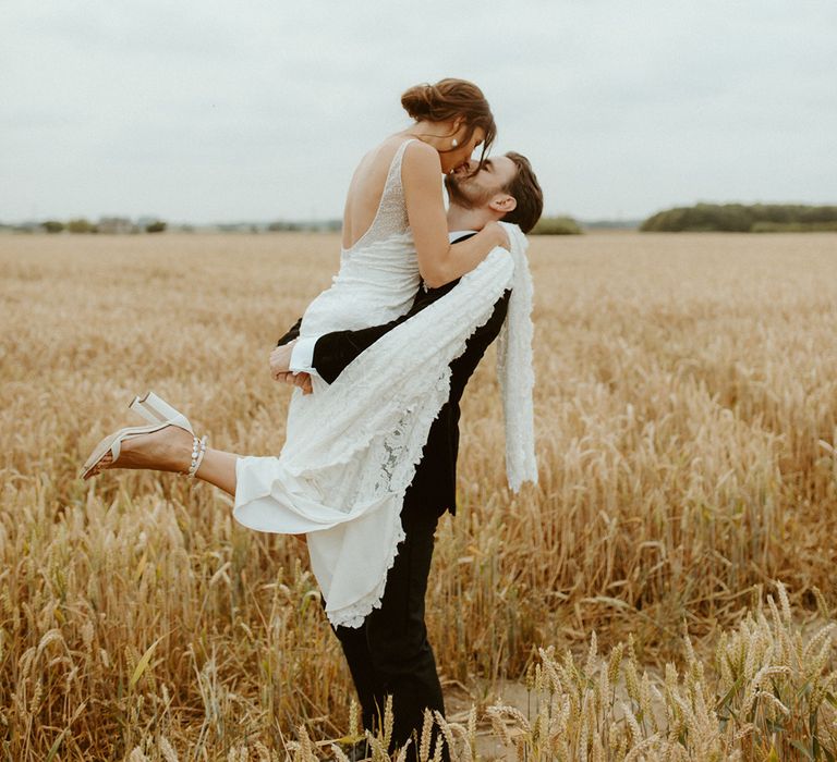 Bride and groom in a field as the groom lifts the bride and kisses her 