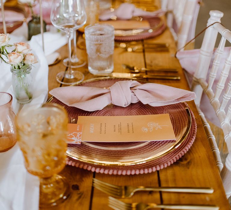 Place setting with pink placemats, gold rimmed plates and glasses, pink napkins and yellow on the day wedding stationery 