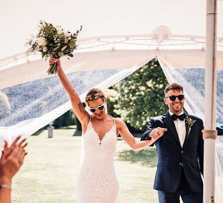 Groom in blue tuxedo with black sunglasses walks in with the bride in lace wedding dress with retro heart sunglasses 