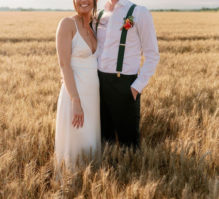 Bride wearing silk satin wedding gown & groom stand in golden fields 