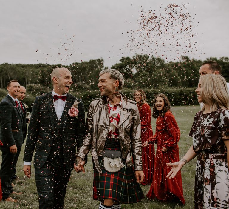 Grooms walk through confetti outdoors as wedding guests surround them as groom wears kilt and gold leather jacket