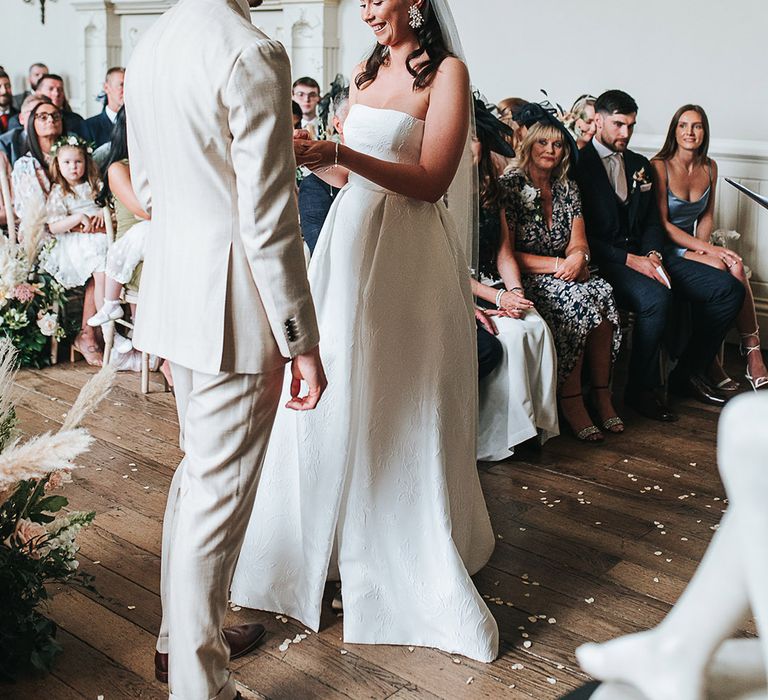 Groom in beige suit stands at the altar as the bride puts a ring on his finger 