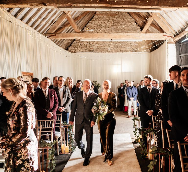 Bride wearing olive velvet wedding dress walks down the aisle in a barn with her father at Bradwell Abbey 