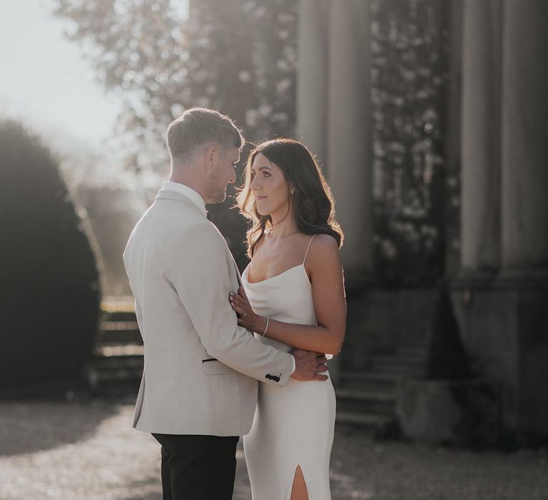 Bride in Alexandra Grecco satin wedding dress with front slit and groom in grey suit jacket gaze at each other 