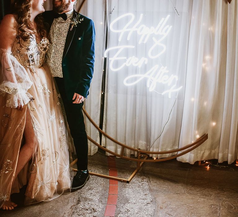 Bride & groom stand beside neon wedding sign that hangs within moon shaped stand in front of curtain 