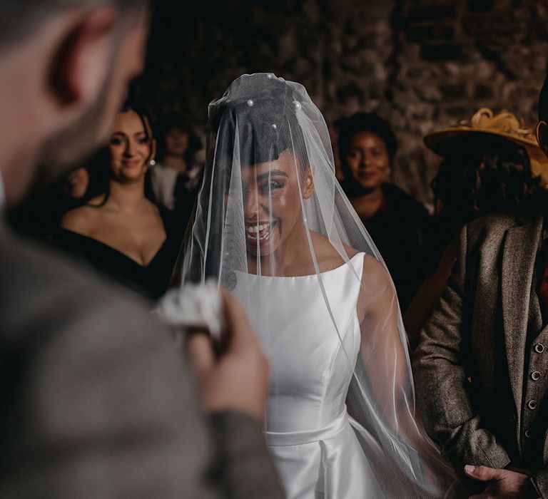 Black bride wears satin Sassi Holford wedding dress with halter-neck style and veil across her face during Christian wedding ceremony