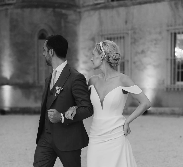 Bride wearing Suzanne Neville wedding dress with off-the-shoulder detailing walks with her groom outdoors 