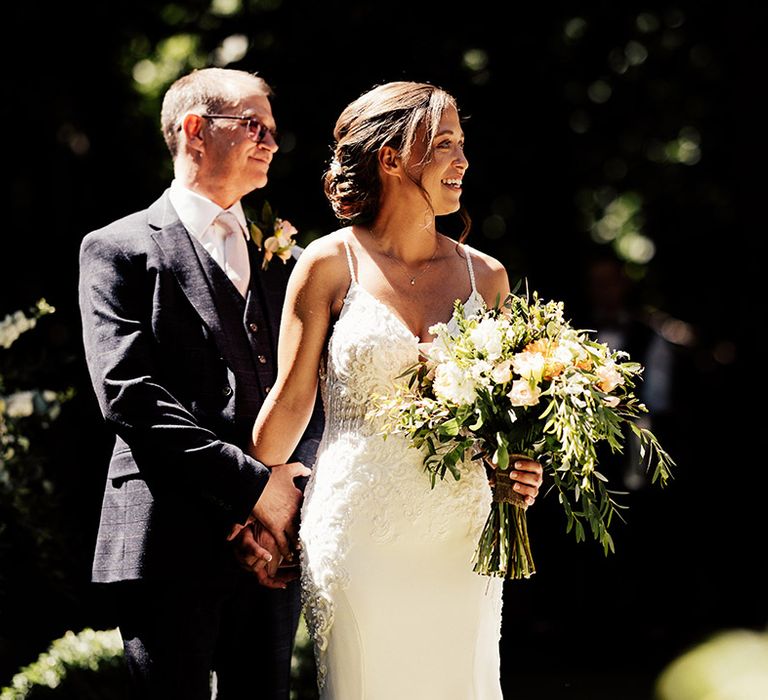 Father of the bride stands with the bride holding her hand as they turn to see the bride walking up the aisle 