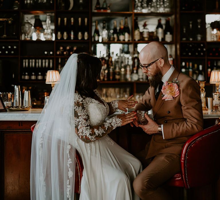 Bride & groom in retro styled brown suit with statement pink floral buttonhole sit at bar drinking whiskey 