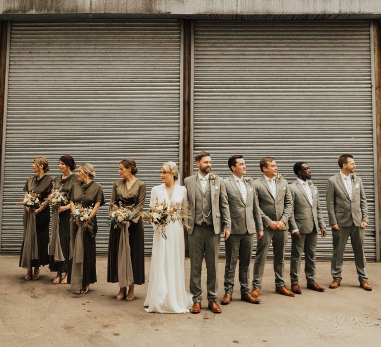 Bridesmaids wear long sleeve green dresses with cowl necks and hold dried floral bouquets whilst stood beside groomsmen in three piece suits