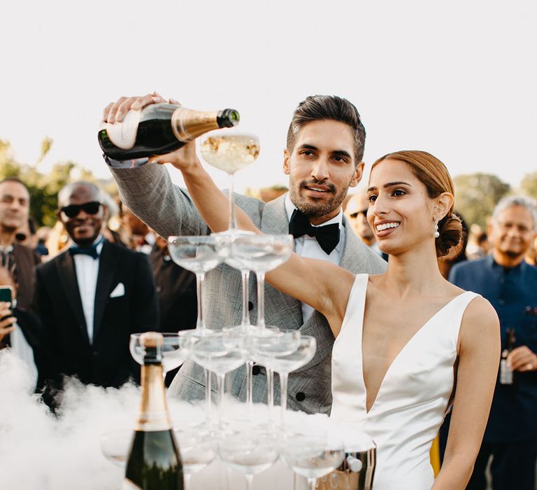 Groom in black tie and his bride pour champagne into champagne tower as wedding guests watch 