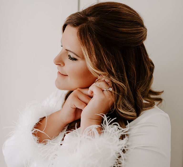 Bride wears silk pyjamas in white complete with fluffy cuffs as she puts in her earrings