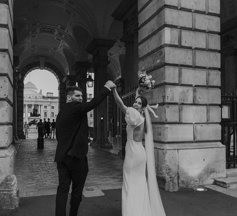 Bride in modern wedding dress with open back detail and long veil holding neutral toned floral bouquet with groom in dark tux holding hands