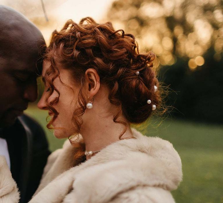 Bride in light fur coat with pearl hair accessories and matching pearl earrings and groom in black tux embracing underneath clear umbrella at Pepper Arden Hall